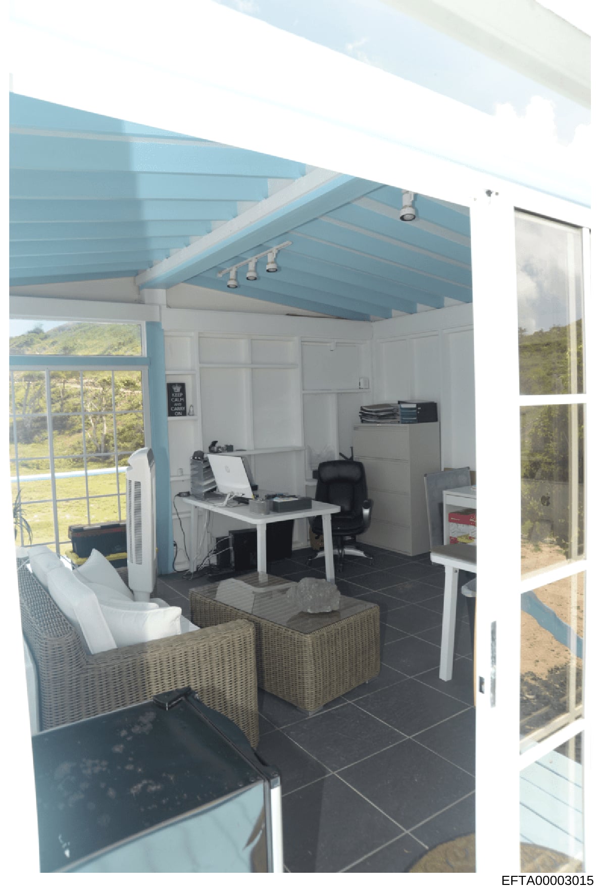 This is a photograph of a contemporary home office or studio space featuring a distinctive blue beam ceiling, work desk with computer equipment, comfortable seating area with woven furniture, and large windows overlooking natural landscape. The space