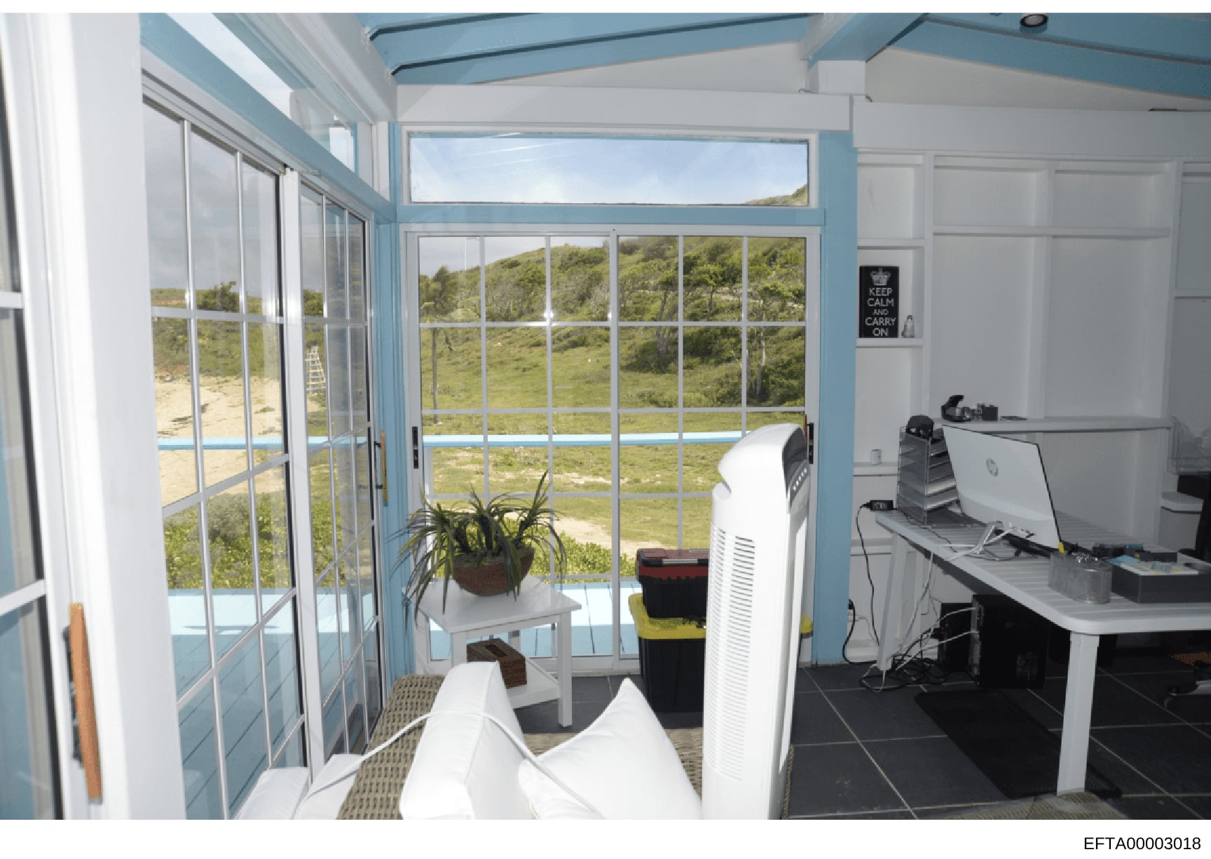 This photograph shows an interior room with extensive windows overlooking a coastal landscape with green hills and water visible in the background. The room appears to be a modern office or study space with computer equipment, an air conditioning uni
