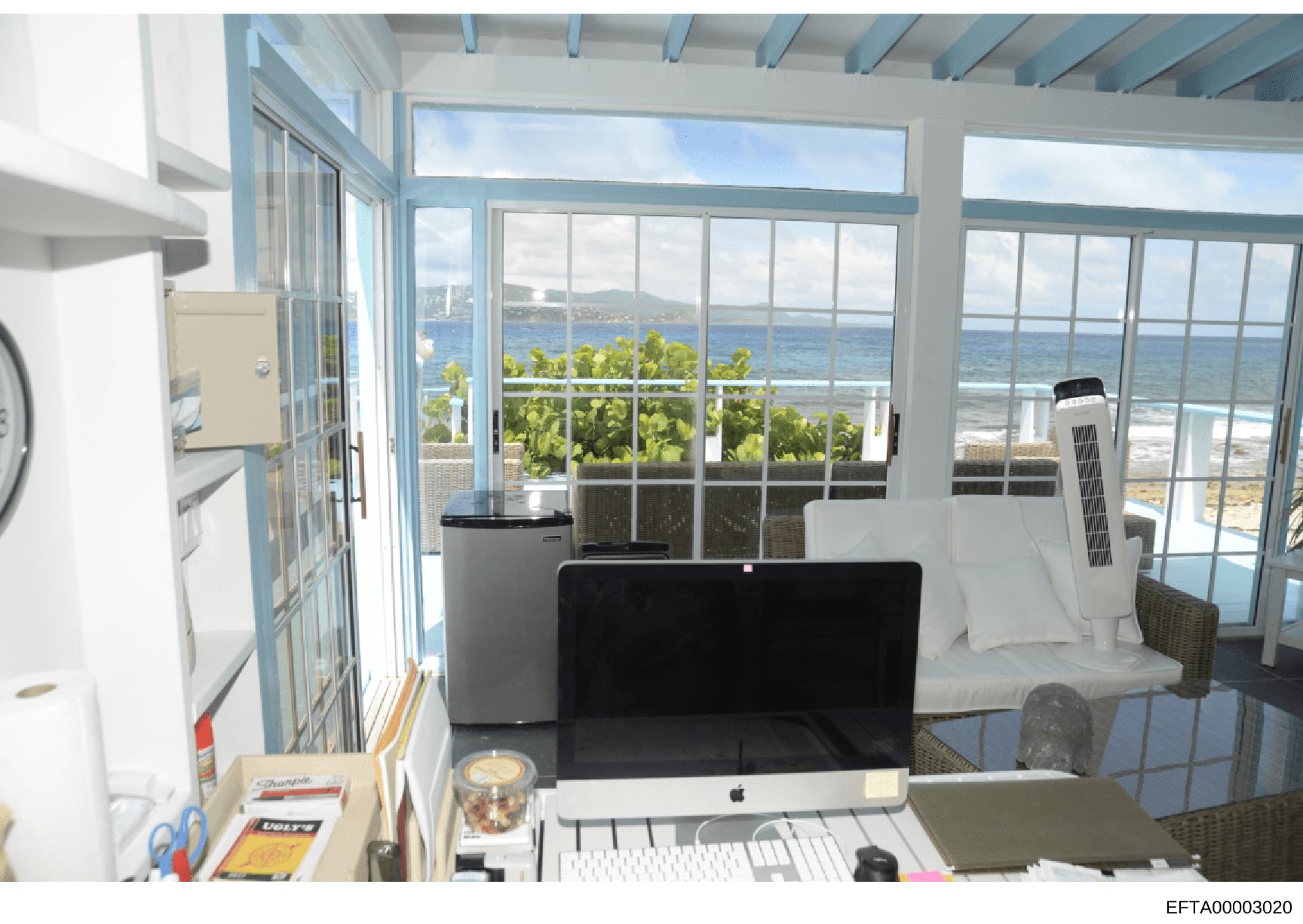 This photograph shows an interior office or workspace in a beachfront property, featuring floor-to-ceiling windows with panoramic ocean views, modern furnishings, and coastal landscape visible beyond. The image appears to be documentation of a reside