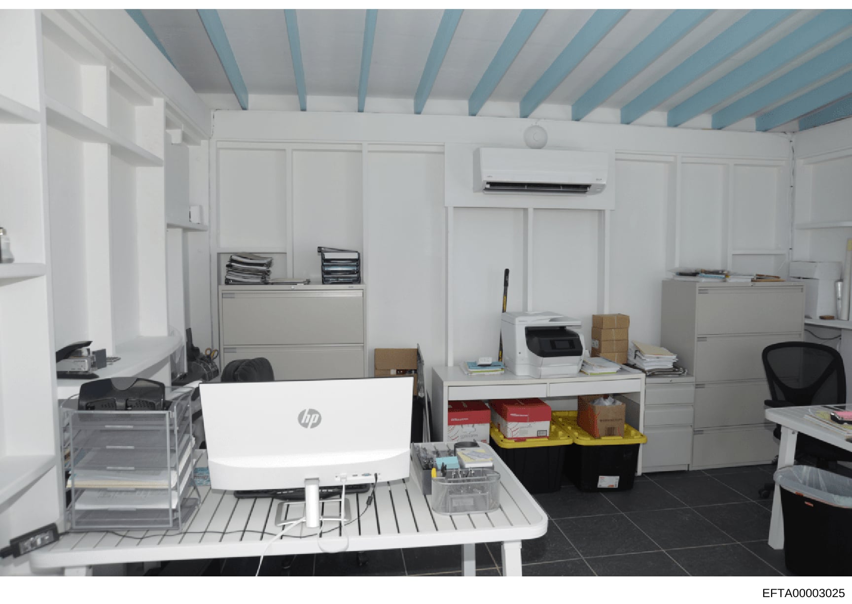 This is a photograph of a modern office space featuring white walls with blue ceiling beams, computer workstations, filing cabinets, and storage units. The room contains an HP laptop, printer, office supplies, and organizational materials in boxes an