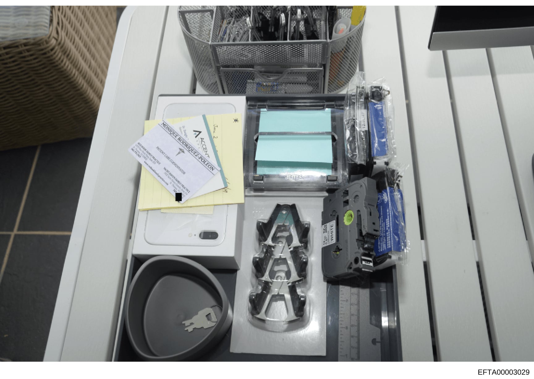 This photograph shows the contents of an office desk drawer containing various office equipment and supplies, including a stapler, tape dispenser, sticky notes with handwritten markings, computer cables, mesh organizer, and other desk accessories. Th
