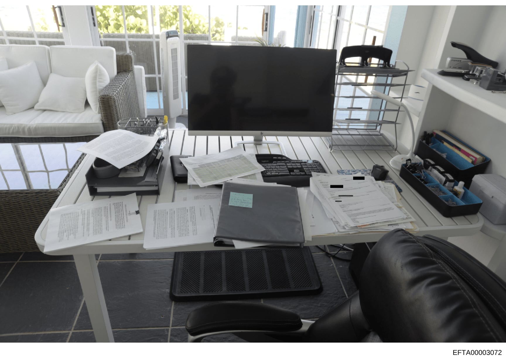 This is a photograph of a bright, modern residential office space containing a desk with scattered documents, a desktop computer setup, and office supplies. The room features large windows, contemporary furnishings including a white sofa with cushion