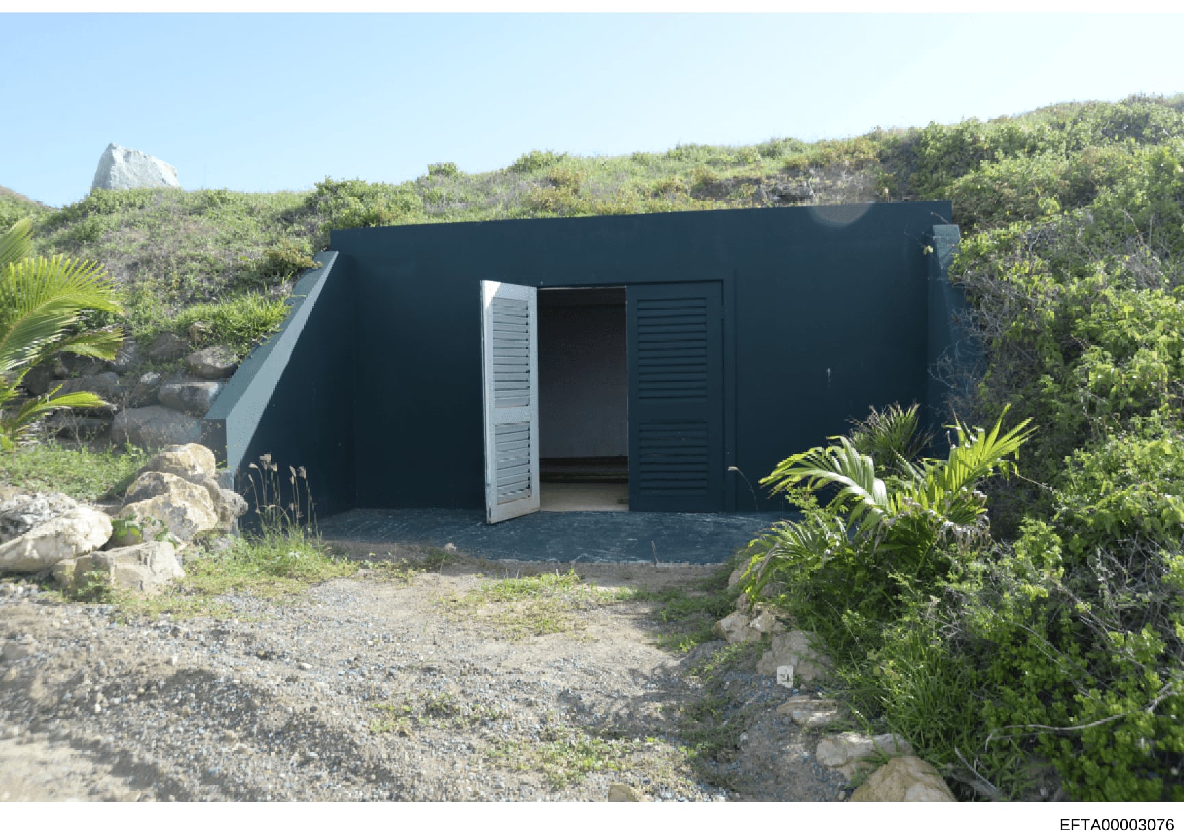 This photograph shows a modern, dark-colored rectangular building or bunker-like structure situated on a remote island location surrounded by dense tropical vegetation and rocky terrain. The building features white louvered doors and appears to be pa