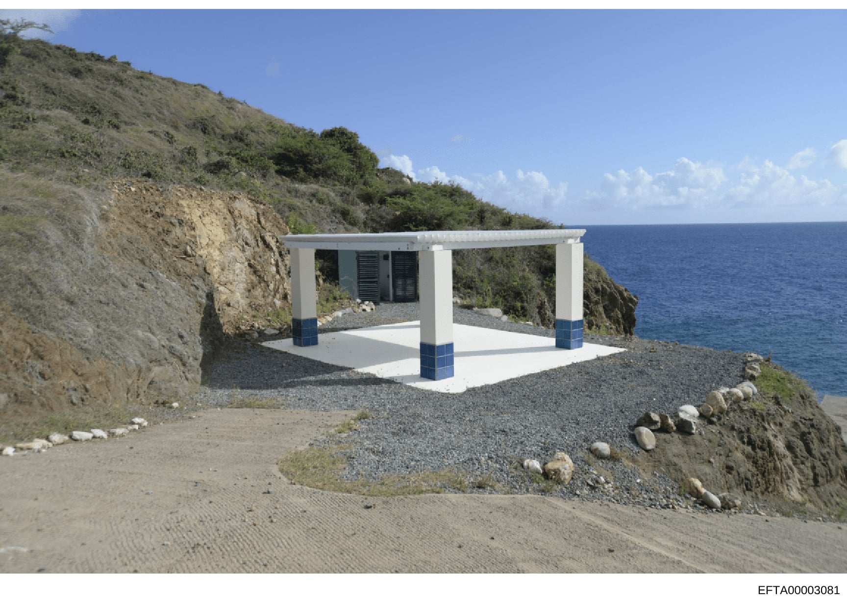 This is a photograph of a modern white structure with blue trim situated on a rocky beach overlooking the ocean. The building appears to be a small pavilion or shelter with a pergola-style roof, louvered windows, and concrete foundation, built into a