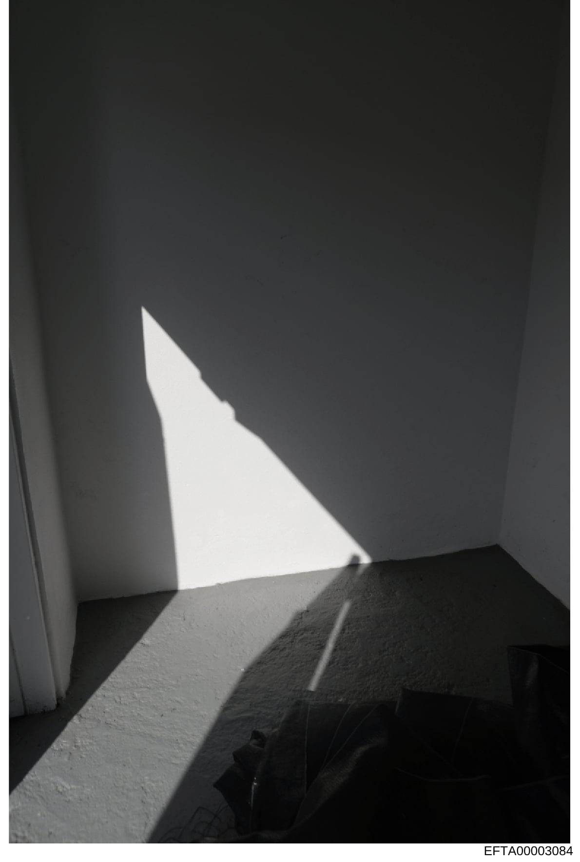 This is a black and white photograph showing an interior corner or architectural space with dramatic natural light streaming through an opening, creating geometric shadows on white and textured walls. The image appears to be a documentary or evidence