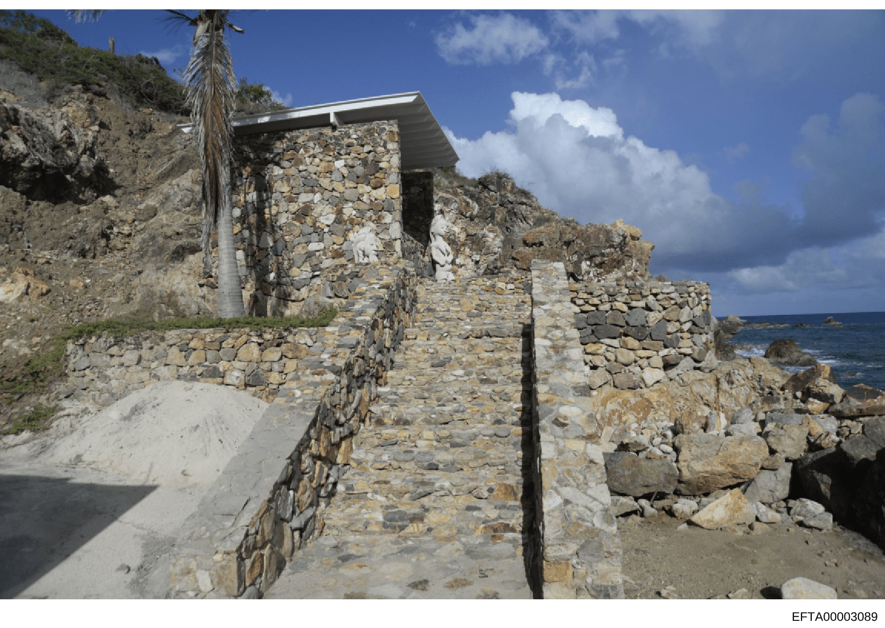 This is a photograph of a stone structure built on a coastal cliff overlooking the ocean. The building features dry-stacked stone walls with a modern metal roof addition and appears to be a residential or outbuilding located on a rocky, elevated terr