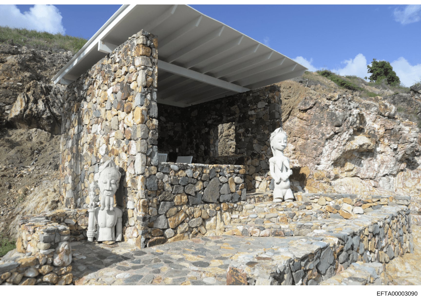 This photograph shows a stone building or structure built into a hillside, featuring two white sculptural figures positioned within and near the structure. The building appears to be constructed from stacked stone with a modern metal roof canopy. The