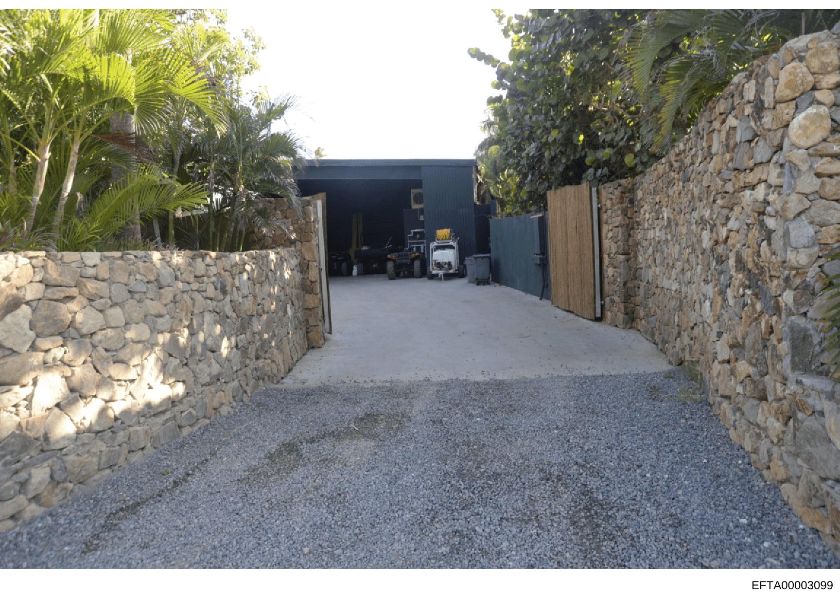 Photograph of a residential property's exterior showing a gated driveway entrance flanked by stone walls, leading to a dark-colored metal outbuilding or garage structure. The image captures the property's access point and security features, with trop