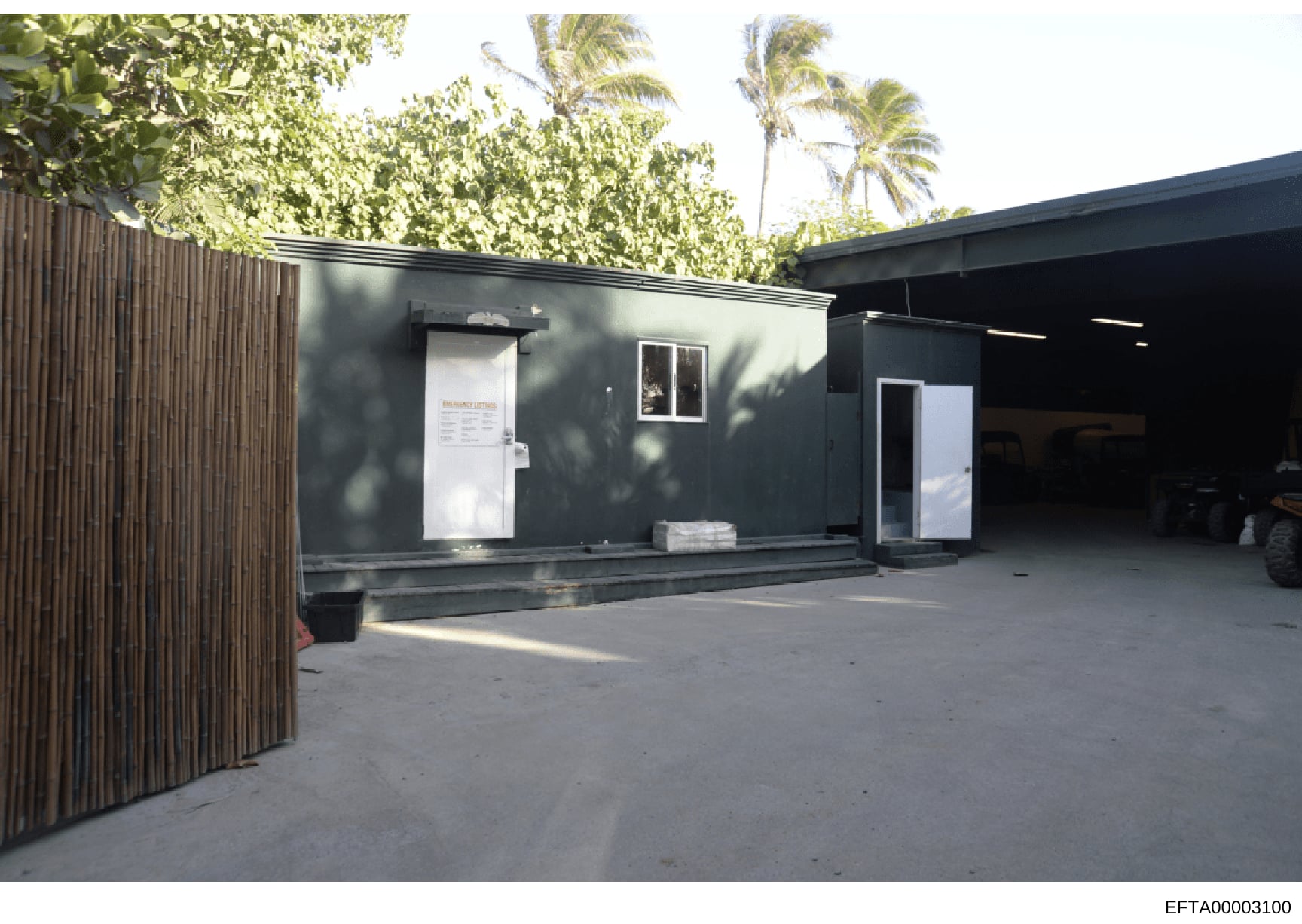 This is an evidence photograph showing the exterior of a modern, dark-colored modular or prefabricated building structure. The building features emergency exit signage visible on a white door, metal framework, and is situated in a tropical setting wi