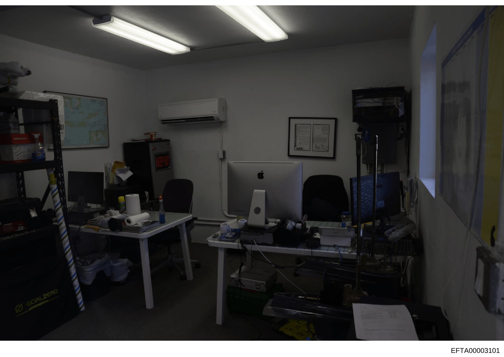 This is an evidence photograph showing an interior office or workspace with multiple computer workstations, including Mac computers, desks, chairs, shelving, and various office equipment. The image appears to be from an investigative search or proper