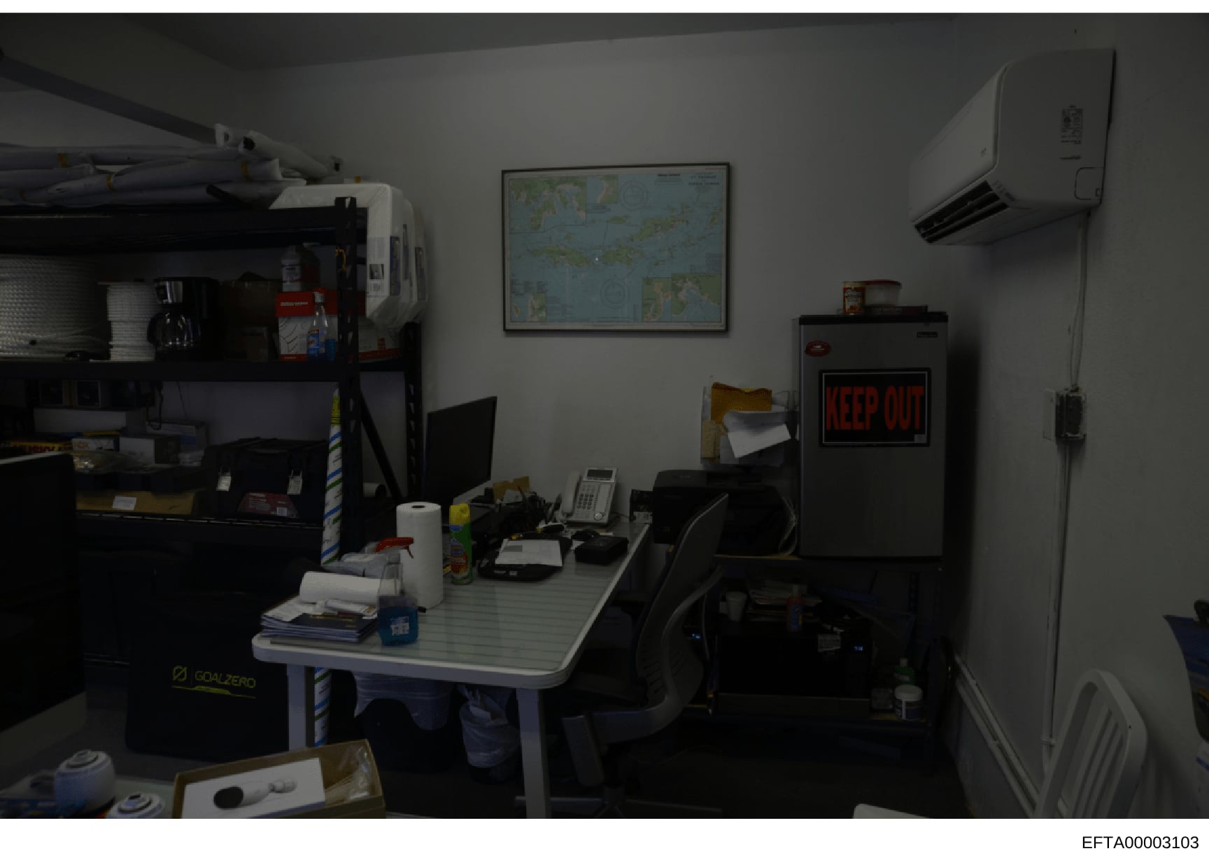This photograph shows the interior of an office or administrative workspace, featuring a desk workstation with computer equipment, storage shelving, and various office supplies. The room contains a wall-mounted map, air conditioning unit, and a 'KEEP