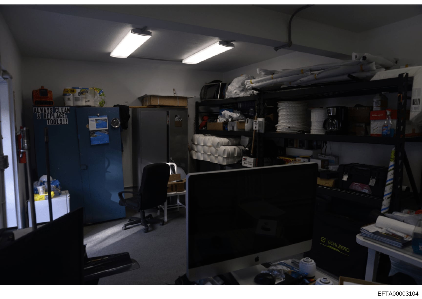 This is a photograph of what appears to be a maintenance, storage, or utility room. The space contains various equipment including refrigeration units, shelving with supplies, cleaning materials, and office furniture. The room is well-lit with fluore
