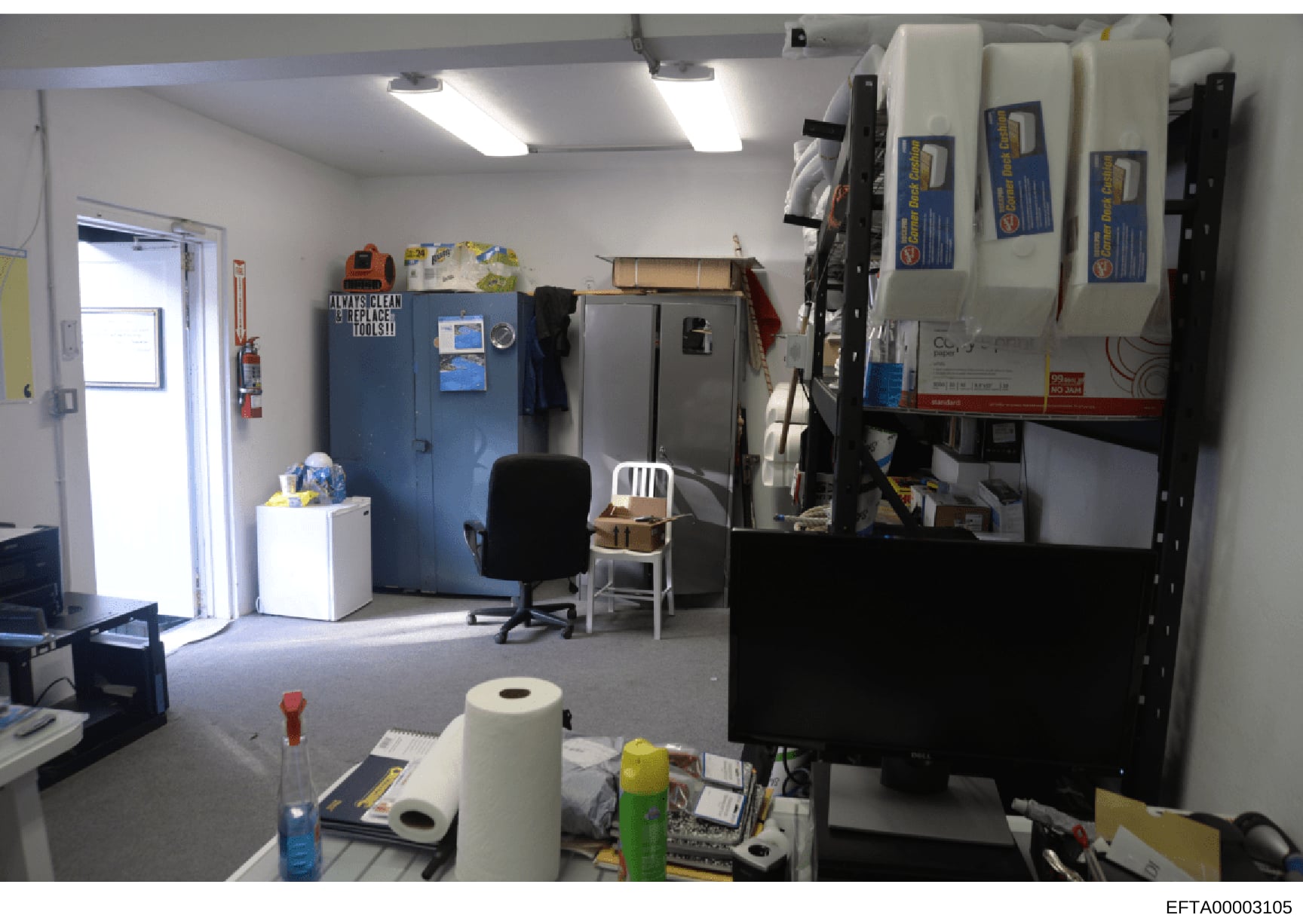 This is a photograph of an interior workspace showing office furniture, storage cabinets, equipment, and supplies. The room appears to be a maintenance, storage, or administrative area with fluorescent lighting, blue storage units, shelving with vari