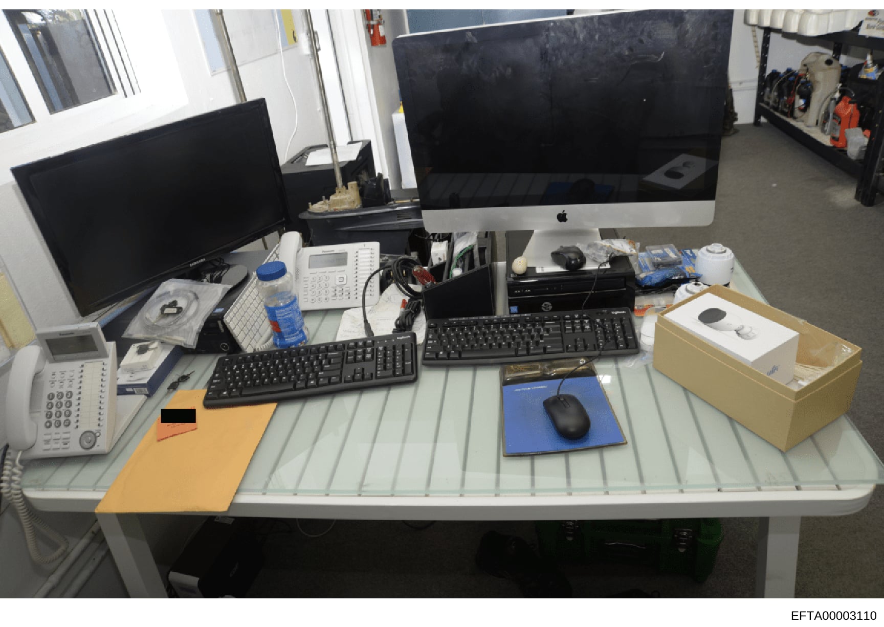 This photograph shows a modern office workspace with dual monitors, computer equipment, telecommunications devices, and various peripherals. The setup appears to be documentation of an office environment, possibly seized or inventoried as part of an