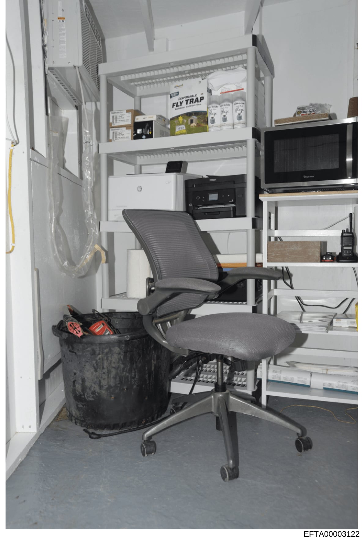 This is a photograph of a small office or work room containing shelving units with various items including office equipment (HP printer, multifunction devices), pest control products (Fly Trap boxes), a microwave, electronics, and miscellaneous suppl