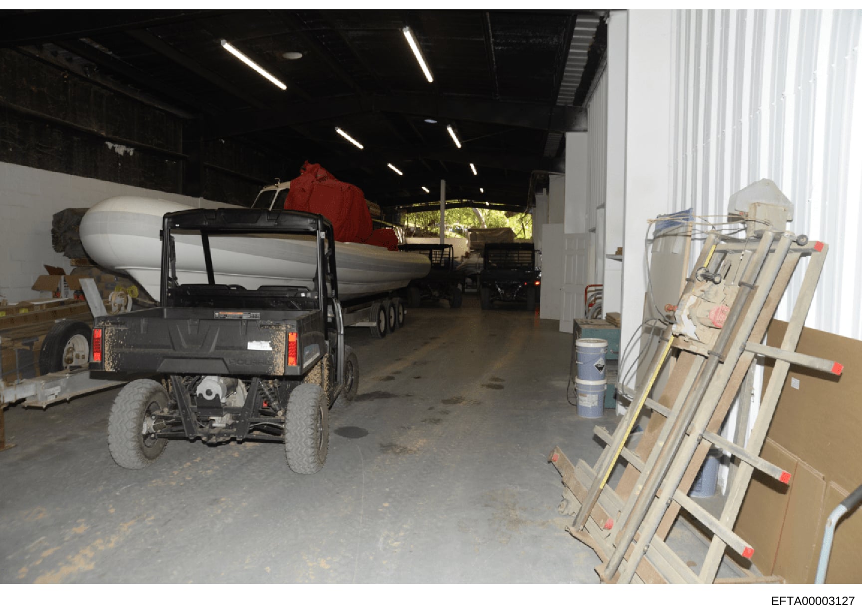 This photograph shows the interior of a large warehouse or storage facility containing multiple vehicles and equipment. The image depicts a black utility vehicle in the foreground, a white tanker truck or vessel in the middle ground, and various cons