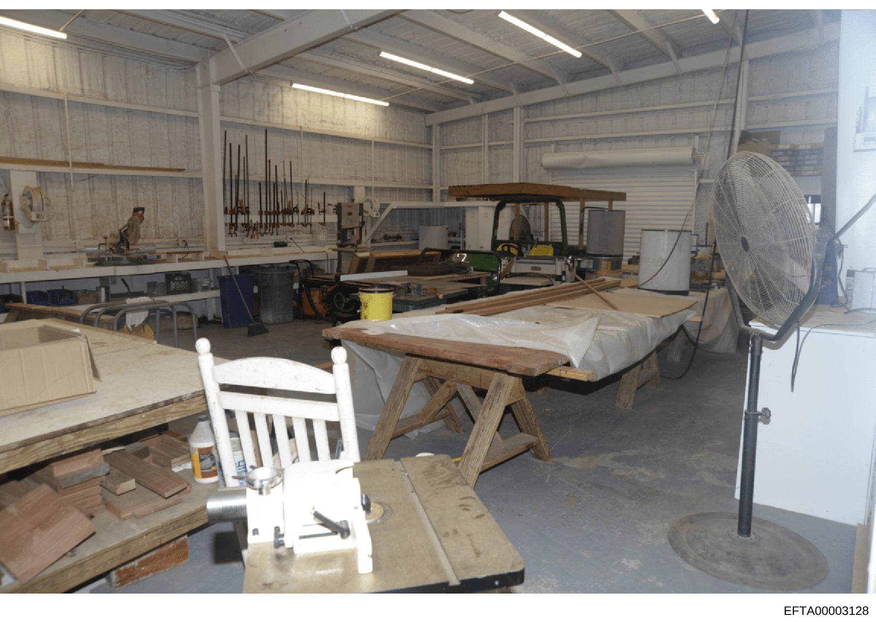This photograph shows a large metal-frame workshop space containing woodworking equipment, materials, and furniture in various stages of completion. The space includes wooden workbenches, a white chair, lumber, power tools, a large industrial fan, an