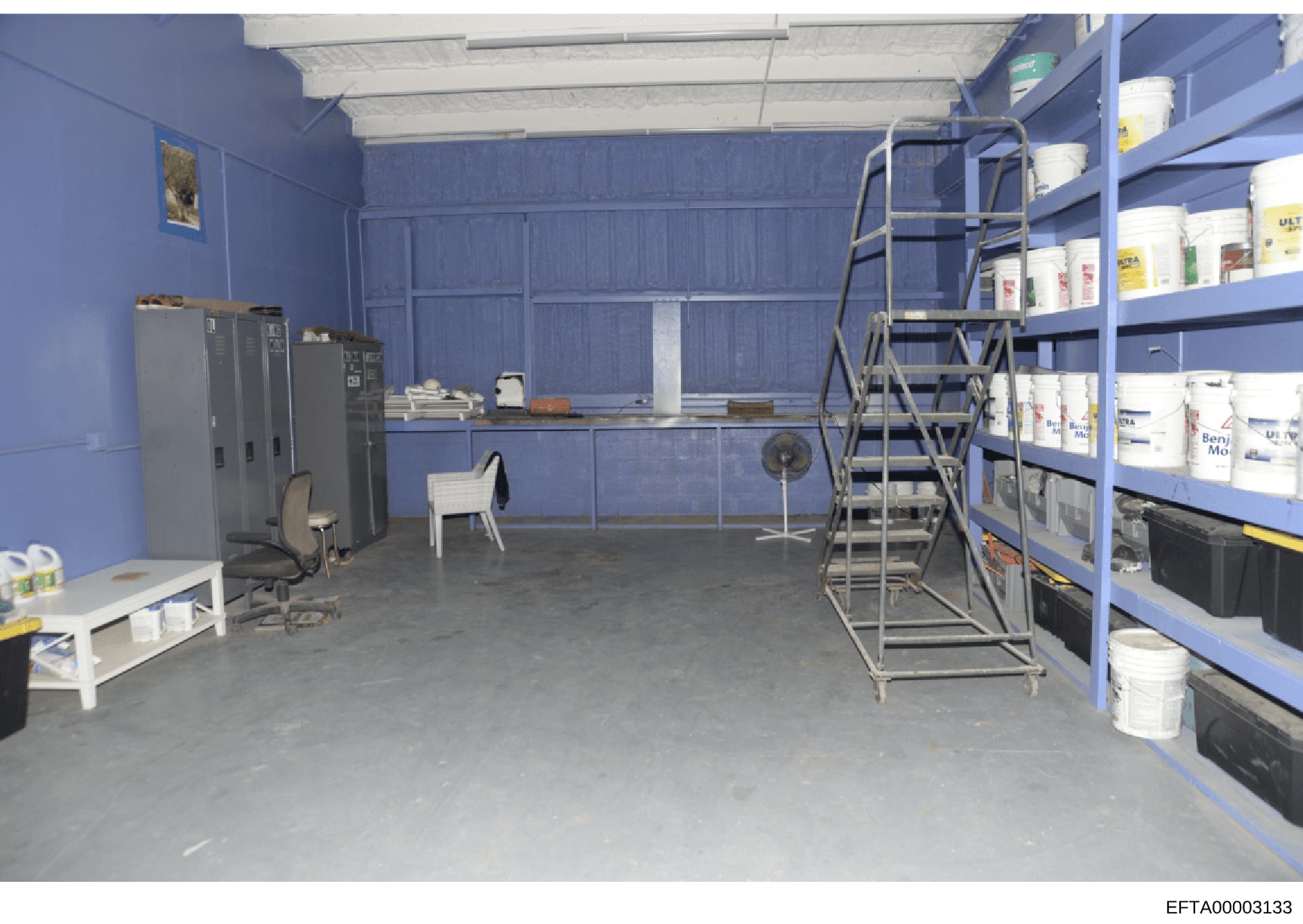 This photograph depicts an industrial storage or warehouse room with bright blue walls and concrete flooring. The space contains metal shelving units stocked with supplies (white buckets and containers), HVAC equipment, office furniture including cha