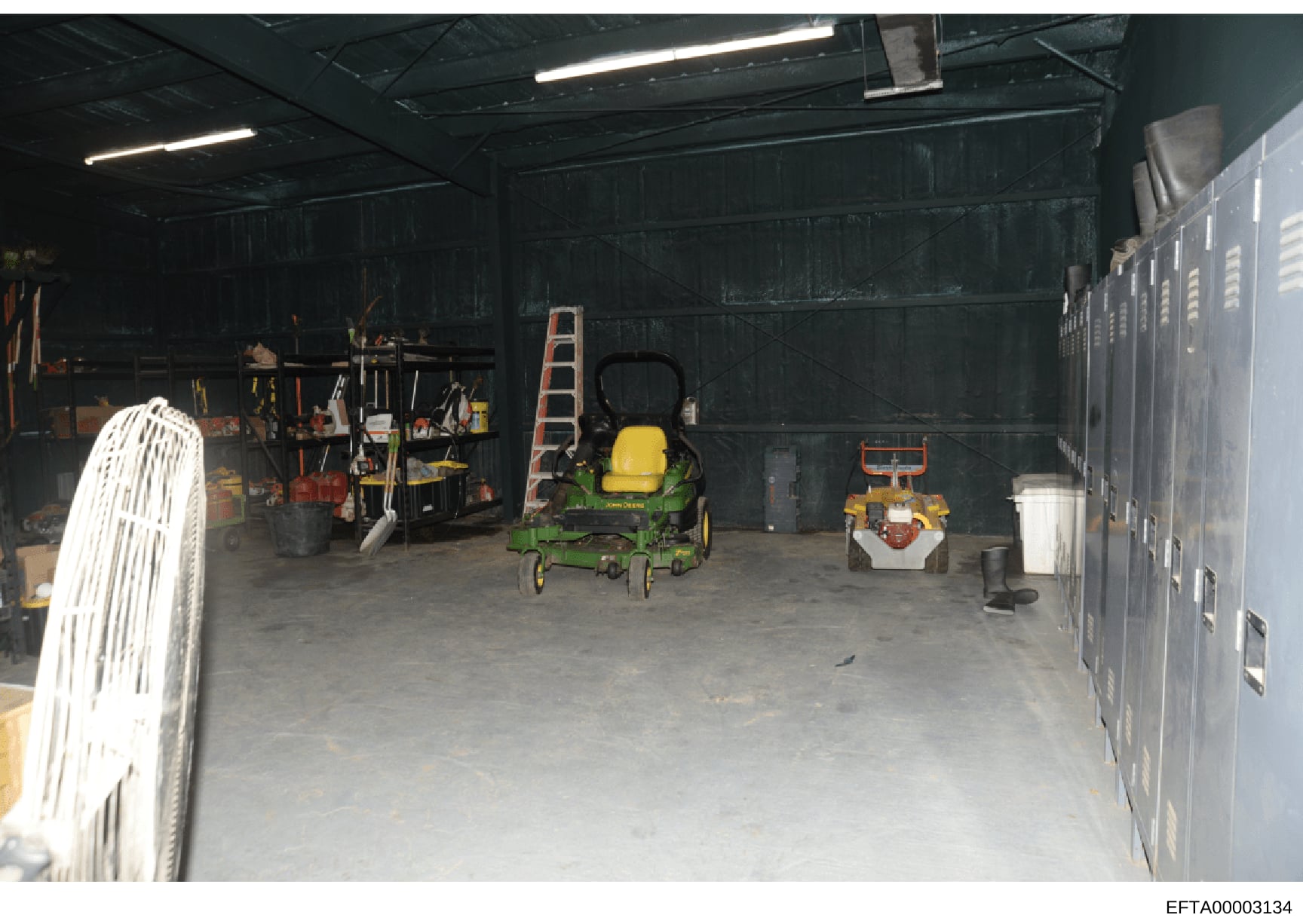 This photograph shows the interior of a storage garage or equipment shed containing various maintenance and landscaping equipment. The space includes a green John Deere zero-turn radius lawn mower, a smaller yellow and red mower, ladders, tools, and