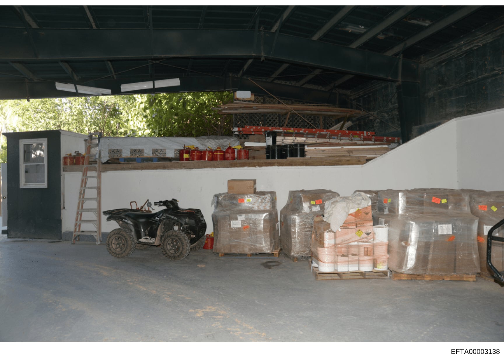 This photograph shows the interior of a warehouse or storage facility containing multiple palletized items wrapped in plastic with inventory tags, a black ATV/four-wheeler vehicle, construction materials, and storage shelving. The image appears to be