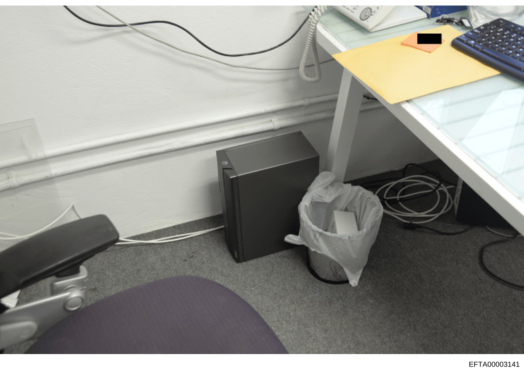 This is an evidence photograph showing an office workspace with a desk, computer tower, keyboard, and other office equipment. A trash bin is visible under the desk. The photo appears to be part of a property search or evidence collection documenting 