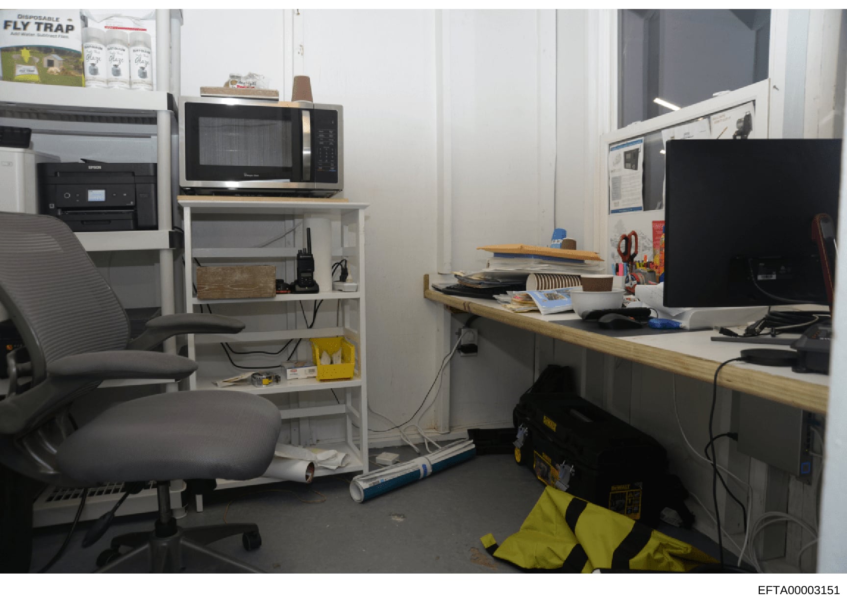 This is a photograph of a small office or workspace containing computer equipment, shelving, a desk, and various storage items. The room appears to be a work area with a microwave, multiple monitors, filing materials, and office supplies. The photo w