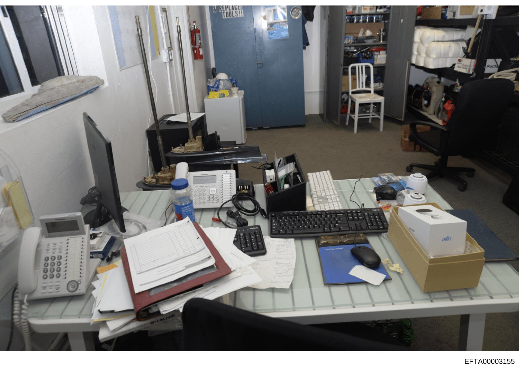 This is an evidence photograph showing an office or workspace, likely from an Epstein-related property search or seizure. The image captures a desk area with computer equipment, telecommunications devices, filing materials, and various storage items.