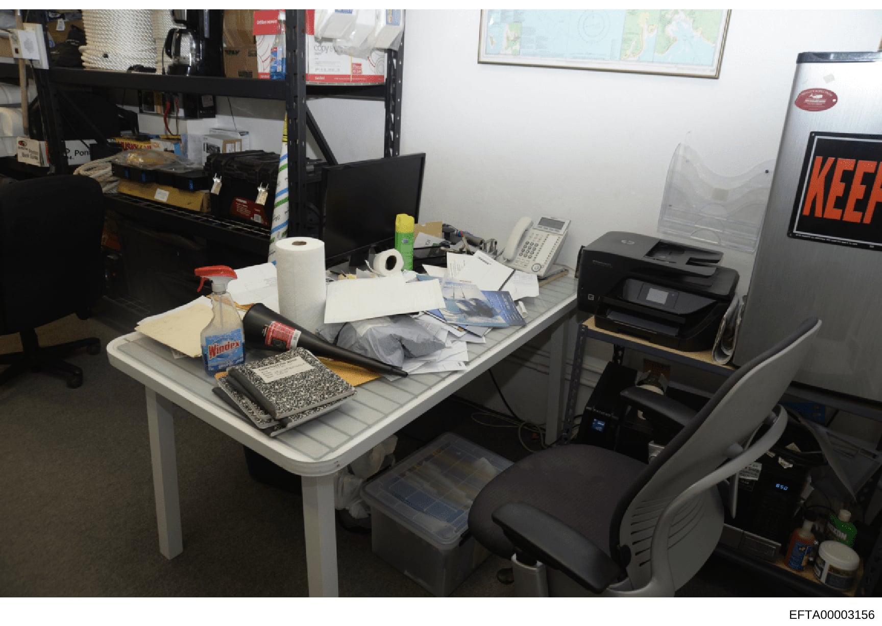 This is an evidence photograph of an office workspace showing a desk with various items including computer equipment, papers, office supplies, and personal effects. The cluttered desk and surrounding area suggest this may be documentation of a locati