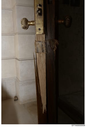 This is an evidence photograph showing the interior detail of a wooden door frame and brass door hardware, likely from one of Epstein's properties. The image captures the door's wooden frame with visible wear and deterioration, brass ornamental door 