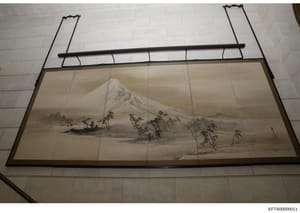 This photograph documents a framed piece of Asian artwork, likely a painting or print on silk or paper depicting a misty mountain landscape with a snow-capped peak, trees, and water. The artwork is mounted in a traditional frame with horizontal suppo