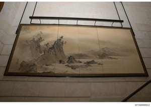 This photograph shows a framed traditional Asian landscape artwork installed on a ceiling, featuring a monochromatic mountainous scene with water, rocks, and sparse vegetation in classical ink painting style. The artwork is mounted in a black-framed 