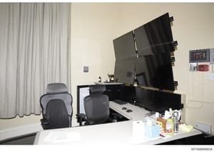 This is a photograph of a modern office space featuring multiple computer monitors mounted on a wall, ergonomic office chairs, and what appears to be a security or monitoring station. The setup includes surveillance or control room-style equipment wi