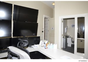 This is a photograph of a modern office room, likely part of an Epstein property, showing a workspace with multiple large computer monitors mounted on the wall, ergonomic office furniture, and adjoining rooms visible through open doorways. The space 