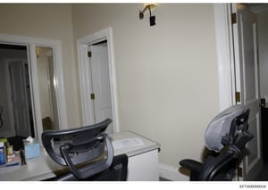 This is a photograph of an interior room, likely an office or bedroom space, showing multiple white doors with brass hardware, beige walls, a desk with office chairs, and basic furnishings. The image appears to be documentation of a property interior