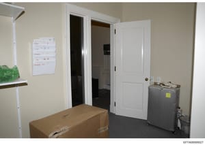 This photograph shows an interior room with organized travel documentation posted on the wall, including labels for various locations (GSJ, LSJ, Zorro, PB, Paris). A cardboard box and metal suitcase with a yellow tag are visible on the floor. The ima