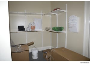 This is an evidence photograph of a storage room or closet containing white metal shelving units with various items. The image shows boxes, bags, a white bucket, green-wrapped items, papers posted on the wall, and other miscellaneous storage contents