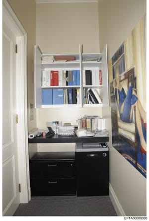 This is a photograph of a compact office closet or storage area containing built-in shelving, file storage, office equipment, and personal items. The space shows organized storage with books, files, and office supplies, along with a black filing cabi