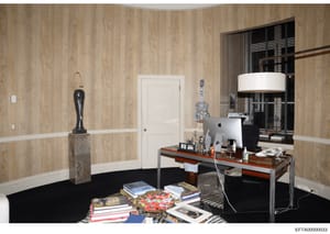 This is a photograph of an upscale office interior, featuring wood-grain wallpapered walls, a modern desk with computer equipment, decorative artwork including a crystalline guitar sculpture, books, and contemporary furnishings. The space appears to 