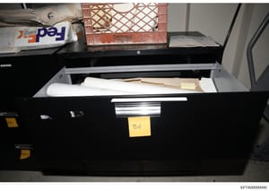 This photograph shows a black evidence storage container or filing drawer that is open, displaying stored documents, rolled blueprints or plans, and various materials. Yellow evidence tags are visible on the exterior, suggesting this is an official e