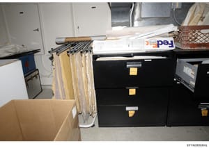 This photograph shows a storage room or evidence facility containing multiple black filing cabinets with yellow identification tags, hanging file folders on a rod system, cardboard boxes, and stacked documents including what appears to be FedEx packa