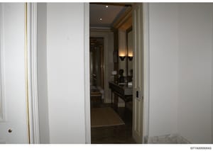 This photograph documents the interior of an upscale bathroom within a residence, showing a double vanity setup with sophisticated finishes, recessed lighting, and decorative wall sconces. The image captures the doorway entrance from an adjoining hal
