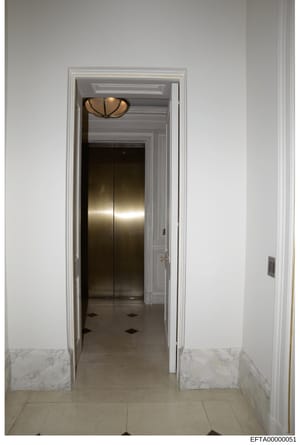 This photograph shows a modern hallway interior with an elevator entrance, marble wainscoting, and decorative ceiling fixture. The image appears to be evidence documentation of a property associated with the investigation, showing architectural detai