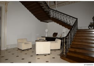 This photograph documents the interior of a luxurious residence, showing a multi-story foyer with an ornate wrought-iron staircase, cream-colored upholstered seating furniture, decorative sculptures, and marble flooring with inlaid pattern work. The 