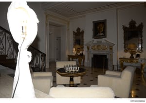 This is a photograph of an ornately decorated formal living room featuring neoclassical and baroque-style furnishings, including gilded mirrors, marble fireplace, decorative sculptures, and cream-colored upholstered seating. A chess set is displayed 
