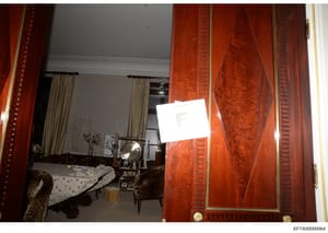 This photograph shows an interior bedroom space featuring prominent red wooden double doors with decorative panels and brass hardware. The room contains a bed with patterned bedding, leopard-print upholstered chairs, cream-colored curtains, and vario