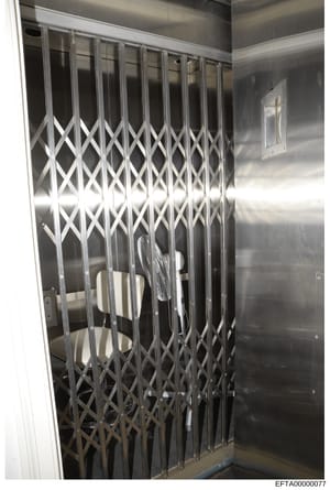 This is a photograph of an interior space featuring a closed accordion-style security gate with a diamond crosshatch pattern in white/light metal. The room has metallic or steel-colored walls and appears to be a secured or restricted area. The image 