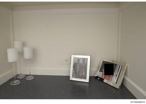 This is an evidence photograph showing the interior of a room with minimal furnishings. The image depicts a dark carpeted floor, cream-colored walls with wall sconce lighting, and three framed photographs placed on the floor against the wall. Two pho