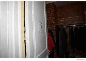 This is an evidence photograph showing an interior closet or dressing room space containing multiple dark-colored garments hanging on a rod, with a red jacket visible among them. The space features white paneling, wooden cabinetry, and wall fixtures.