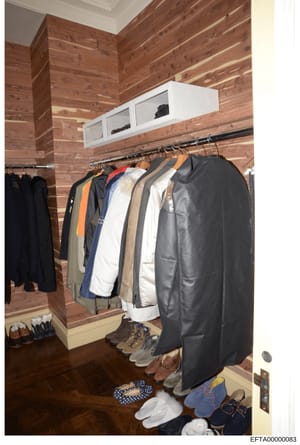 This is a photograph of a wooden closet interior containing multiple garments on hanging rods and various shoes and accessories on the floor. The image appears to be evidence documentation from the investigation, showing the contents of what is likel