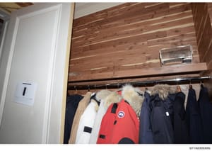 This is an evidence photograph showing the interior of a closet with wooden ceiling beams and walls, containing multiple winter coats and outerwear hanging on a metal rod. The collection includes a red Canada Goose jacket, white, navy, and tan/beige 